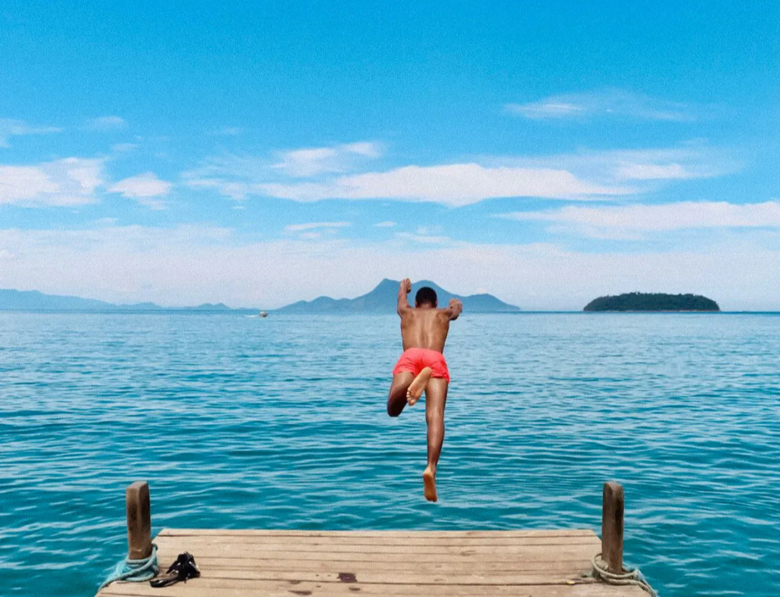A man leaping off a wooden pier toward bright blue sea, with islands on the horizon.