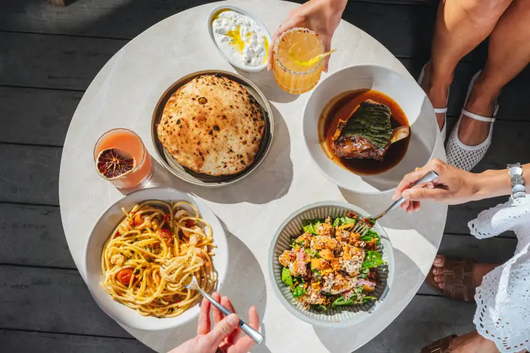 Two people sharing pasta, salad, flatbread and a braised dish at a round table in sunlight, seen from above.
