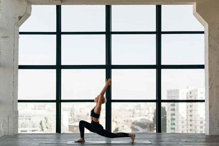 A woman holding a low lunge yoga pose in a bright studio with tall windows.