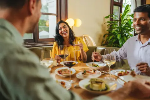 Three friends laughing together over dinner at a restaurant table set with food and wine.
