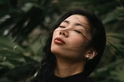 A woman with long dark hair and closed eyes tilts her face up among green leaves.