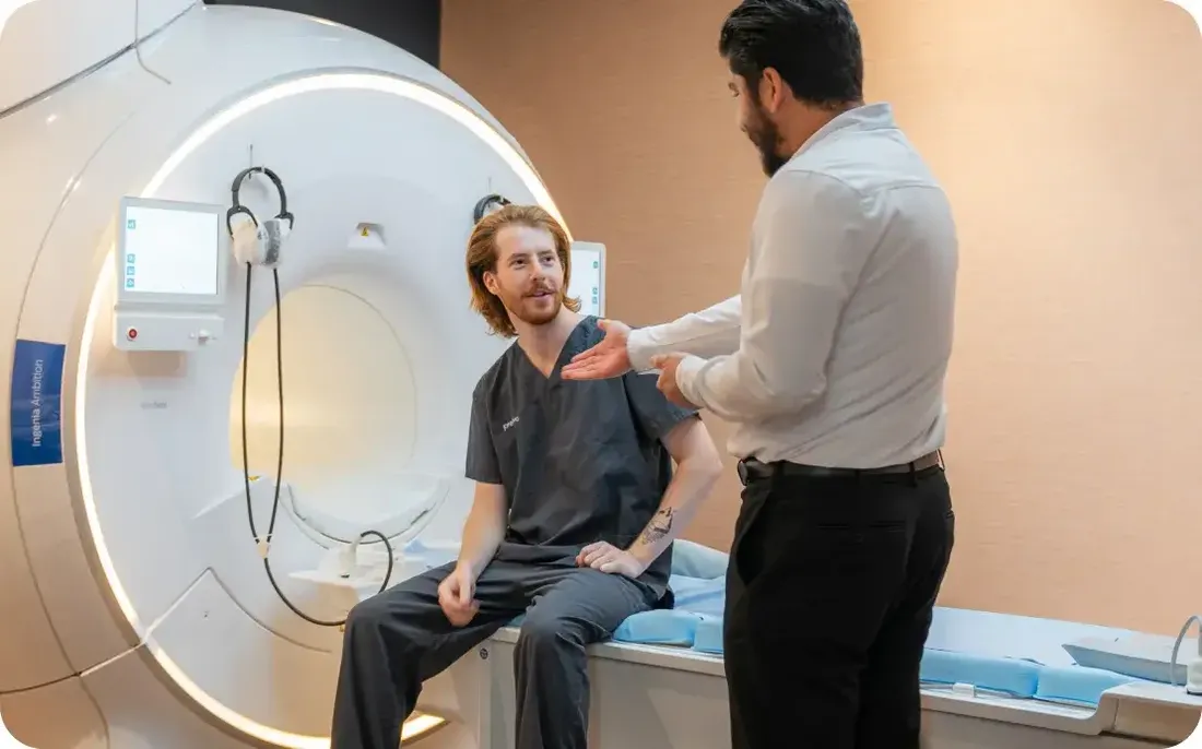 A patient seated on the scanner bed speaking with a staff member in front of a Prenuvo whole-body MRI machine