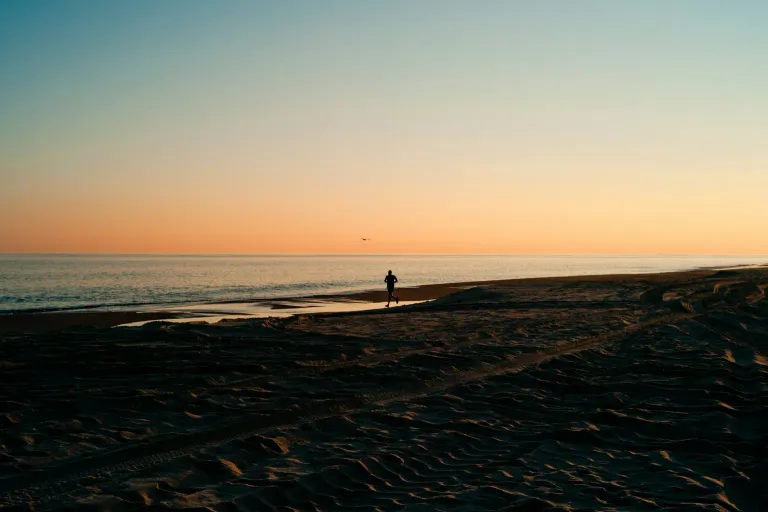 A lone person walking along a quiet beach at golden hour, with a calm sea and an orange sky