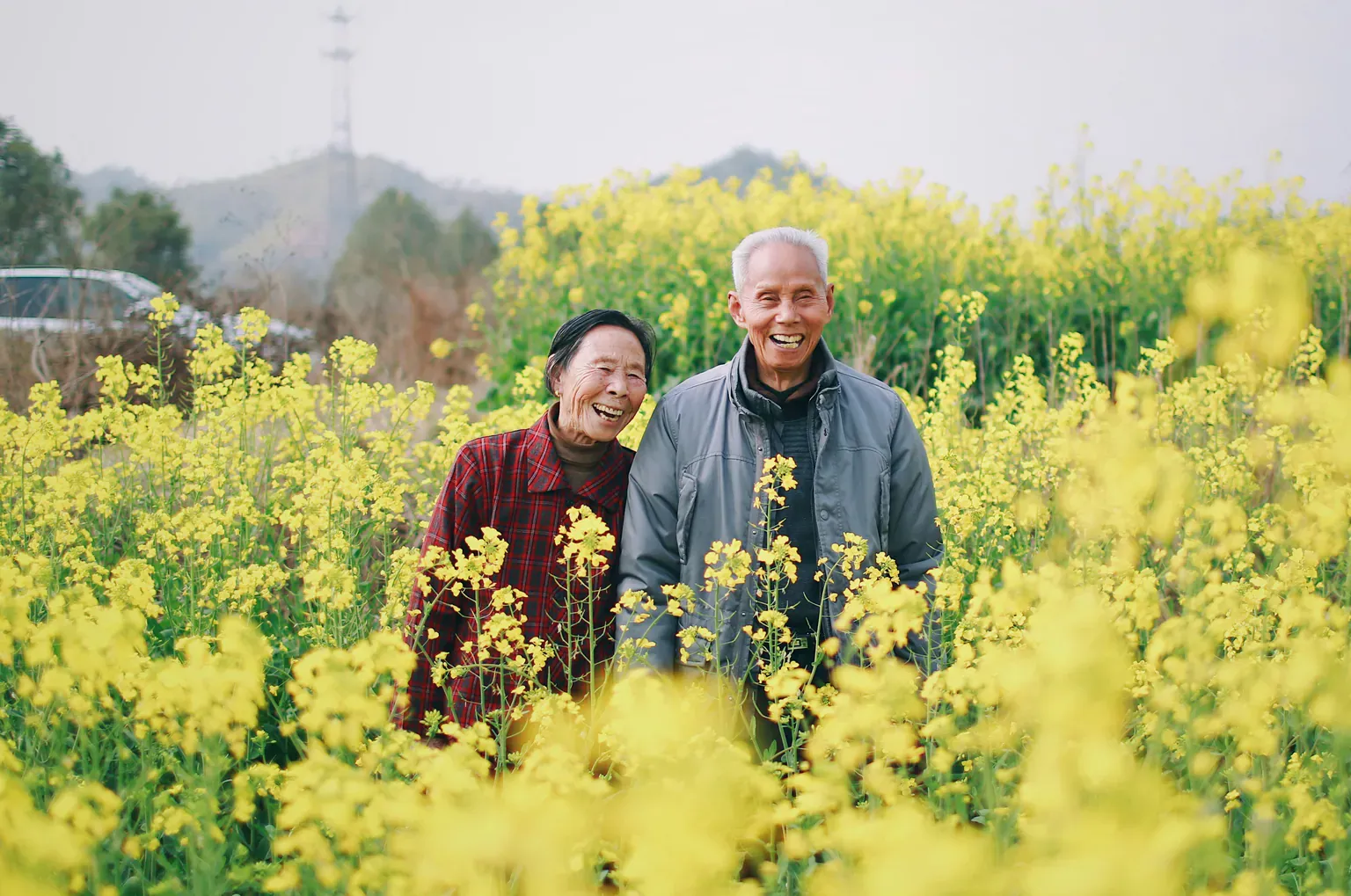 An older couple smile together in a field of yellow flowers.