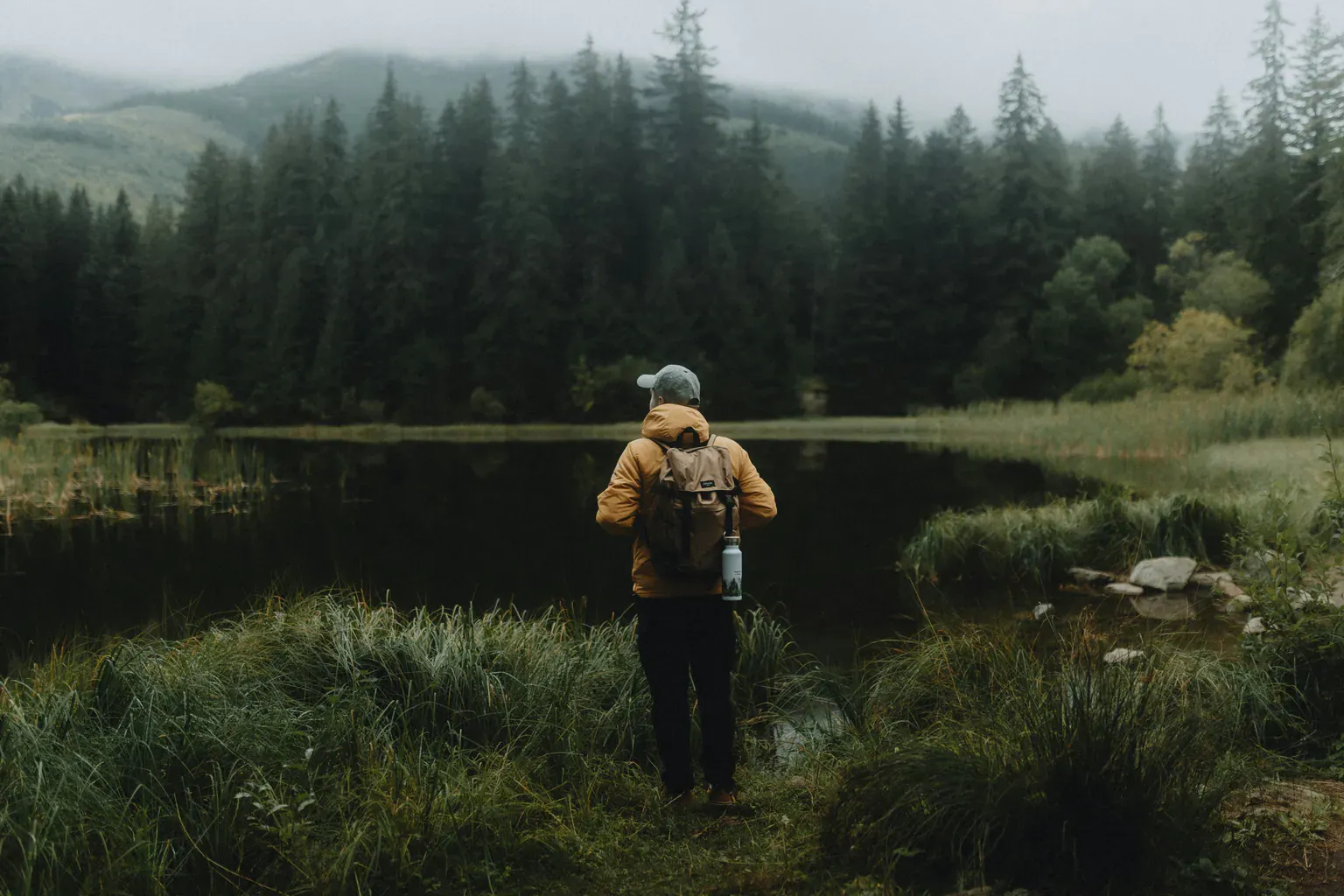 A hiker in a yellow jacket stands at the edge of a still lake below a misty pine forest.