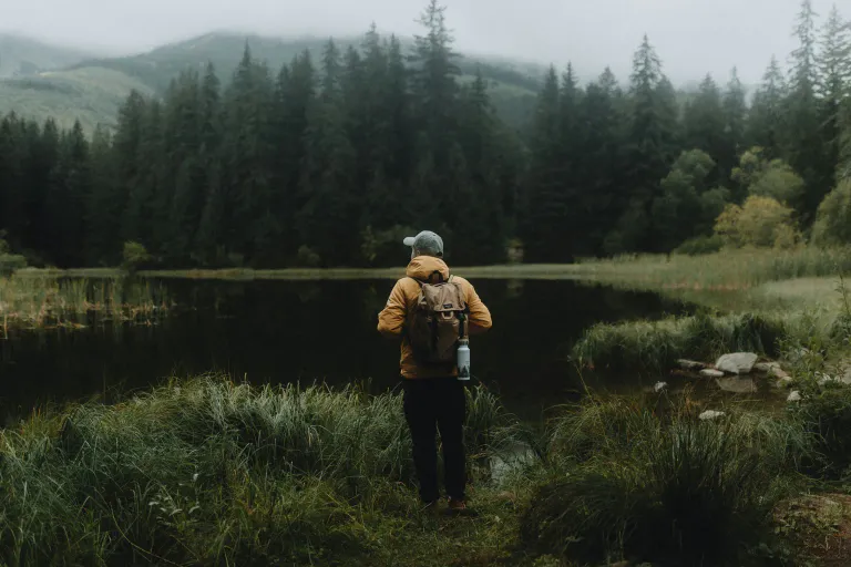 A person with a backpack standing by a dark lake near a pine forest