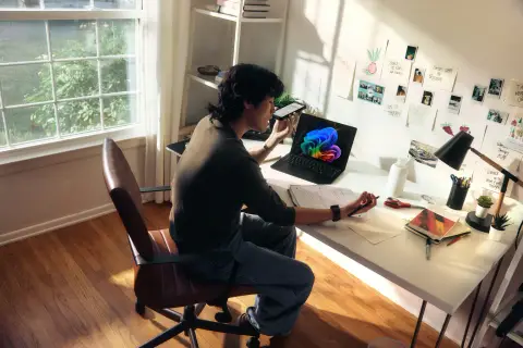 Person at a sunlit desk reading paperwork beside a laptop