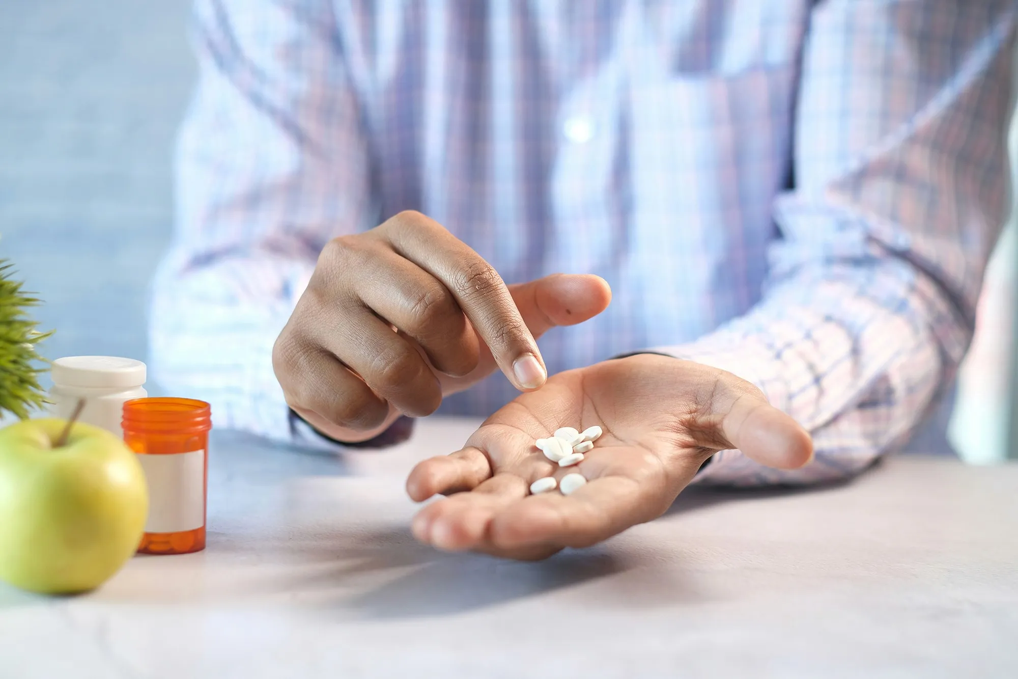 A close-up of a person in a plaid shirt holding several white pills in their hand, with an apple and pill bottles in the background, symbolizing medications or supplements for blood sugar management.