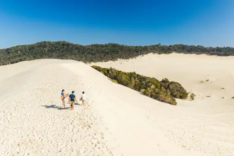 Three people walking up a wide white sand dune beside a green forest under a blue sky.