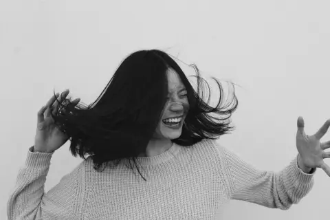 A black and white photo of a smiling woman in a knit sweater, tossing her hair as she laughs.